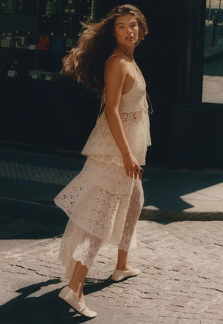 A woman wearing a white lace tiered dress and white flats walks on a cobblestone street in warm sunlight.