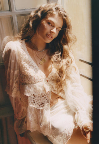 A woman with long wavy light brown hair wearing a sheer cream lace lingerie set with a matching long-sleeve robe